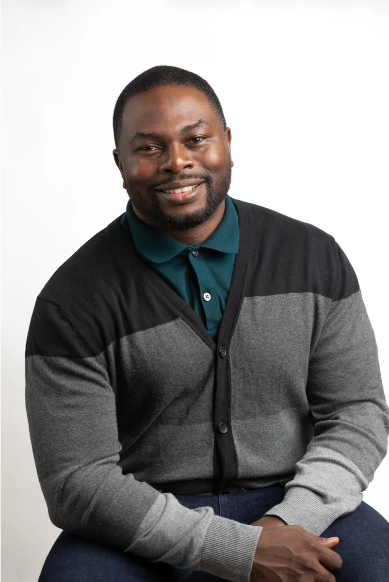 Onome, smiling in a teal polo and gray cardigan.